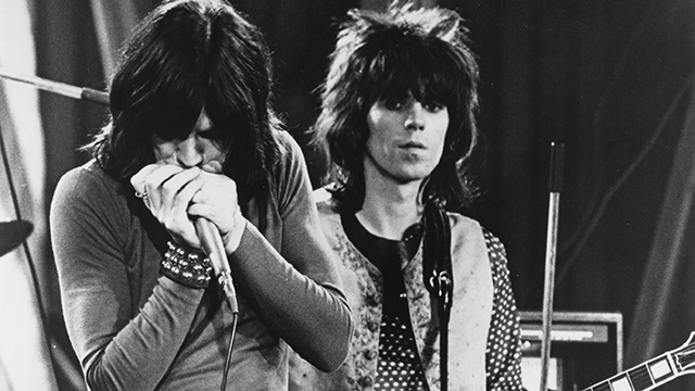 A monochrome picture of two young White men with long hair and rock-style outfits singing and performing.