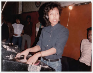 A young woman wearing a headset and looking at someone as she plays a record on a turntable.