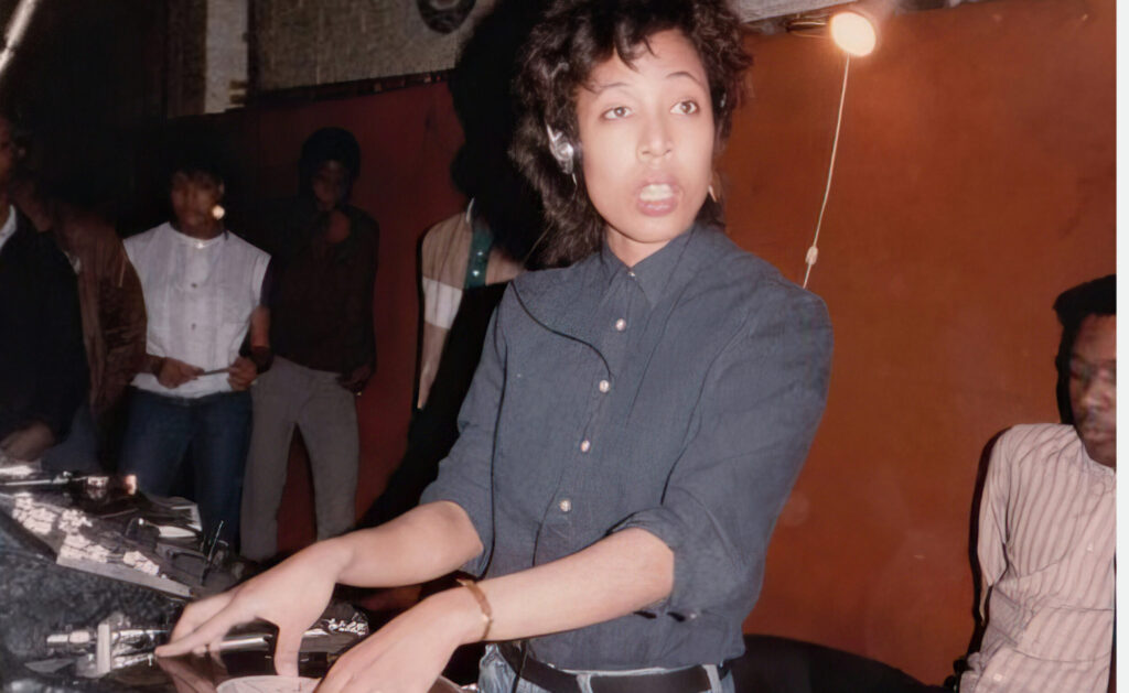 A young woman wearing a headset and looking at someone as she plays a record on a turntable.