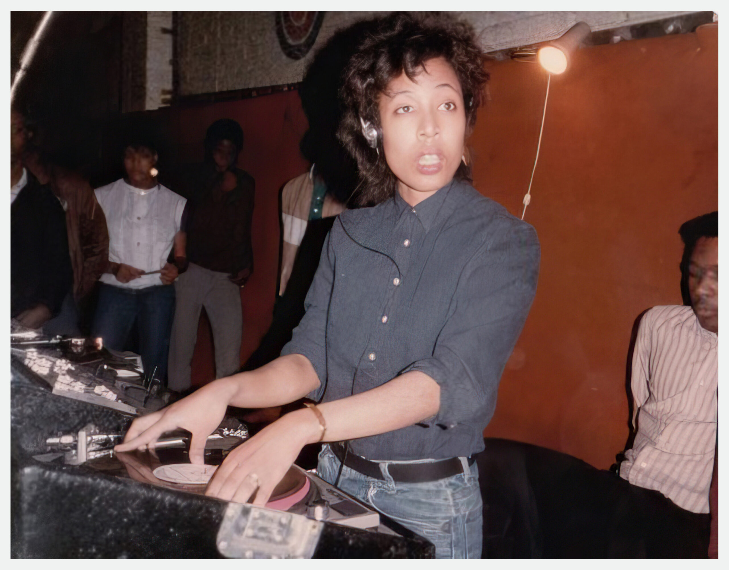 A young woman wearing a headset and looking at someone as she plays a record on a turntable.