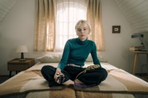 A young woman with short blonde hair wearing green turtle neck and black pants sits on a bed. A pet lizard sits on her lap.