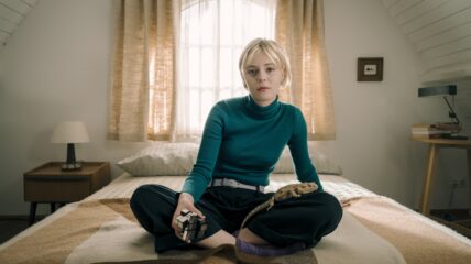A young woman with short blonde hair wearing green turtle neck and black pants sits on a bed. A pet lizard sits on her lap.