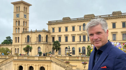 An older White man with silver hair standing in front of a historical building.
