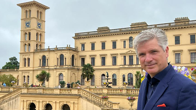 An older White man with silver hair standing in front of a historical building.