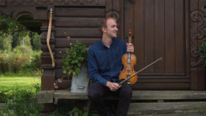 A white man earing a blue shirt and Black pants holds a fiddle