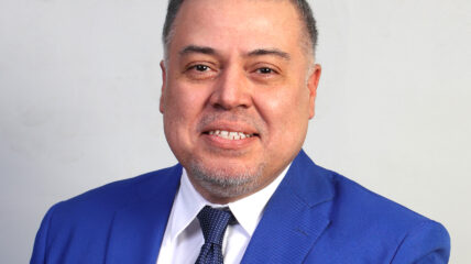 A Latino male with black short hair wearing a blue blazer, white shirt and dark blue tie.