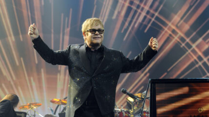 Elton John wearing a sparkly black jacket and black sunglasses standing on stage with his arms raised