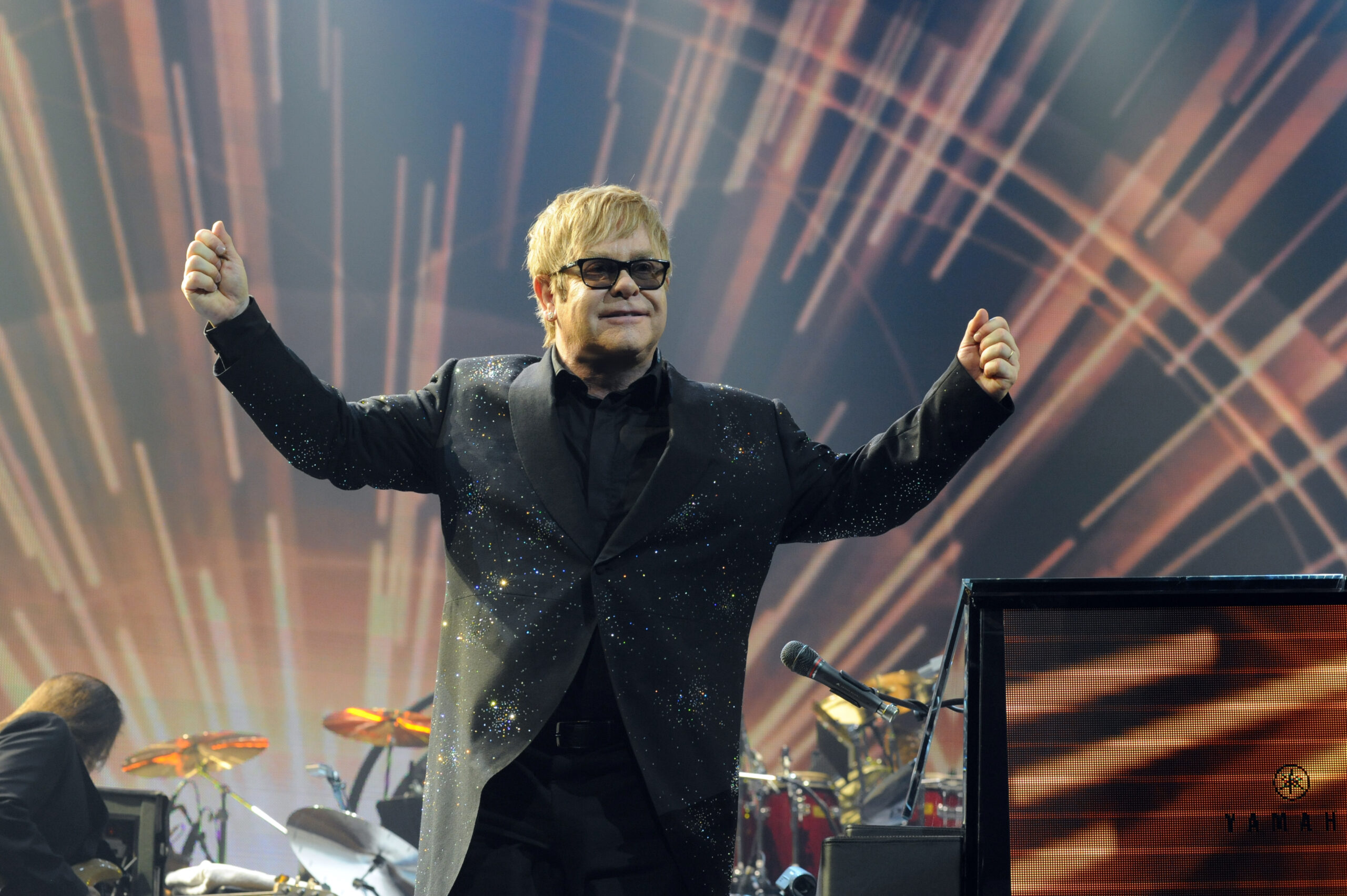 Elton John wearing a sparkly black jacket and black sunglasses standing on stage with his arms raised