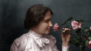 A young Helen Keller wearing a white blouse with a bow at the collar. She is smelling a pink rose.