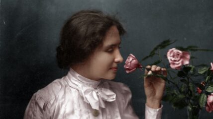 A young Helen Keller wearing a white blouse with a bow at the collar. She is smelling a pink rose.