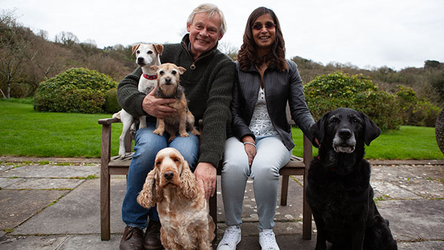 Martin Clunes with two dogs on his lap, sitting with a brown woman who is blind in a park with two more dogs seated both on her left and right.
