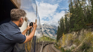 A man leaning outside of a train, clicking a picture via a camera of a scenic view of trees and snowy peaks.
