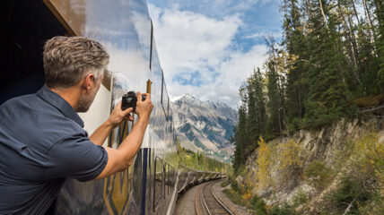 A man leaning outside of a train, clicking a picture via a camera of a scenic view of trees and snowy peaks.