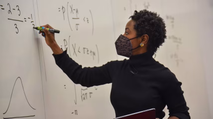 A Black woman with a mask on, writing on a white board
