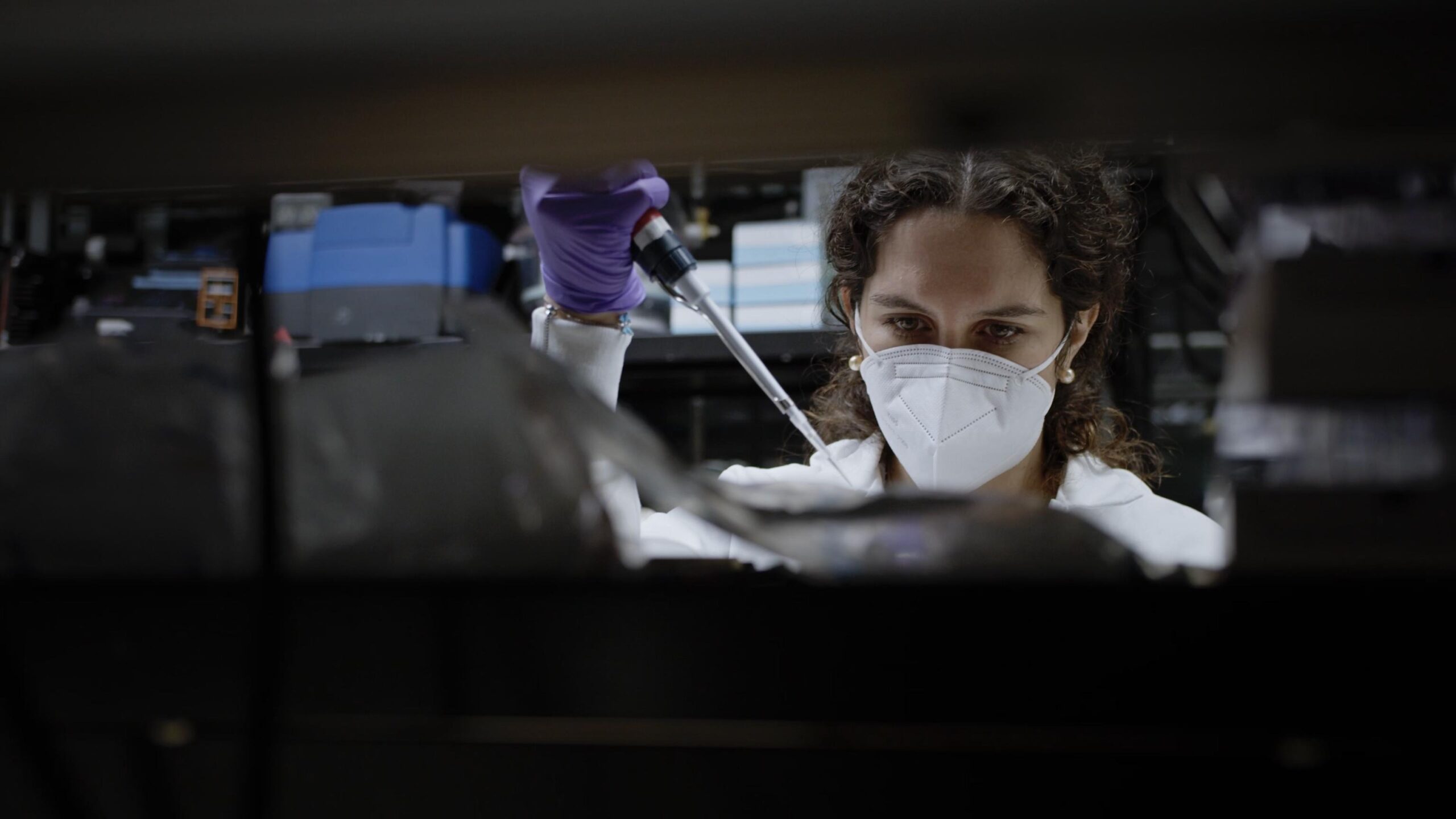 A woman researcher wearing a mask holds a dropper and places contents into a dish.
