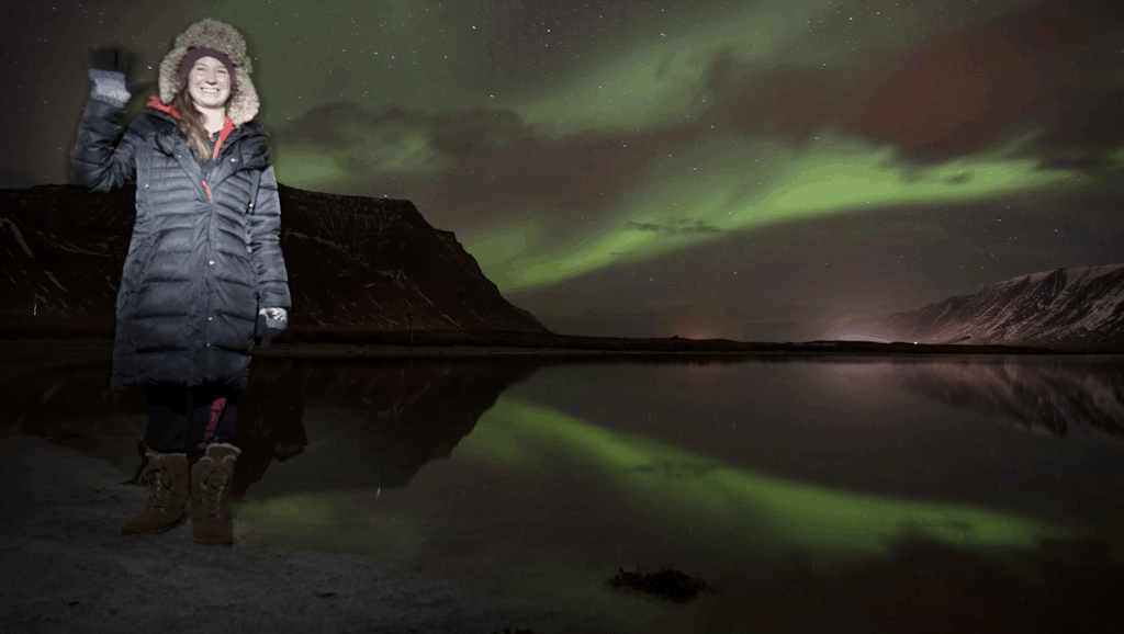 Host Stephanie Gardner standing near o body of water in Flateyri, Iceland at night. She's wearing a Black winter goad and brown boots