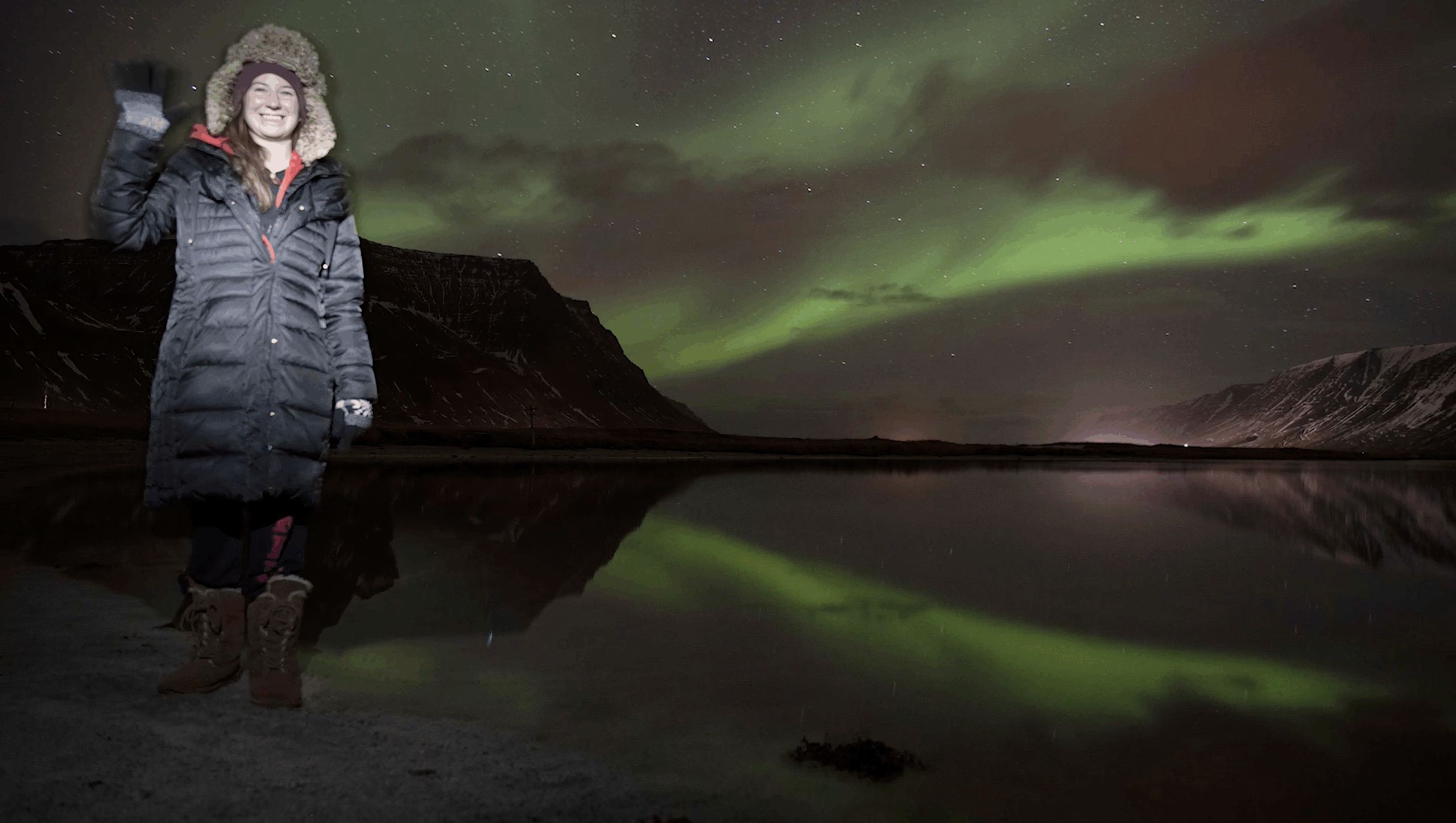 Host Stephanie Gardner standing near o body of water in Flateyri, Iceland at night. She's wearing a Black winter goad and brown boots