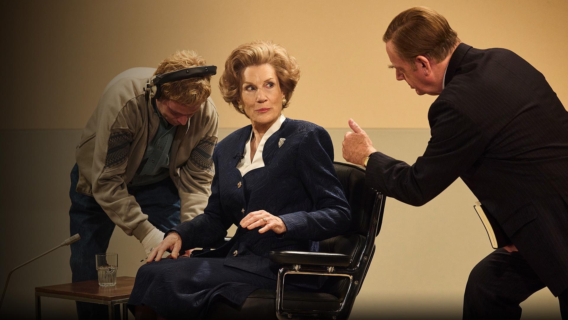 An actress playing Prime Minister Margaret Thatcher wearing a blue suit sits in a chair getting ready to go on air for an interview.