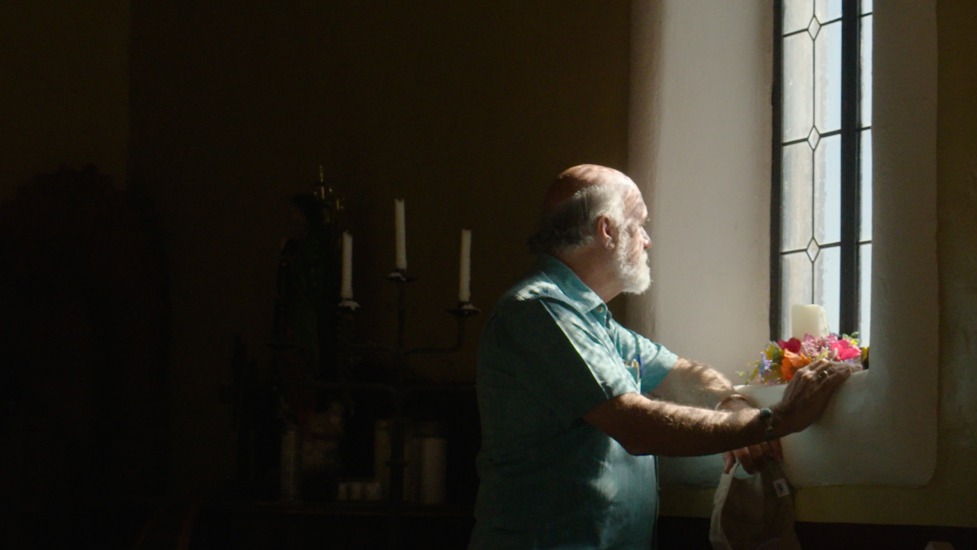 An older white man with gray hair stands in front of a window/