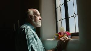 An older white man with gray hair stands in front of a window/