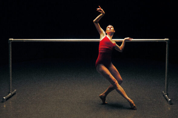A ballerina wearing a red body suite posing a bar.