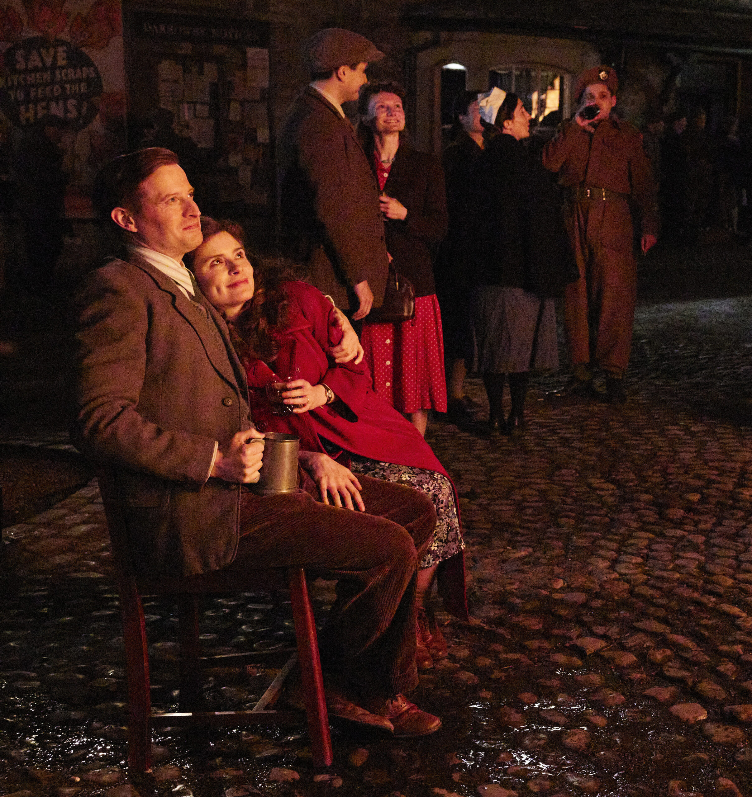 A white man in a brown suit sits next to a white woman in a red jacket. It's evening and they they are looking up a lit sky.