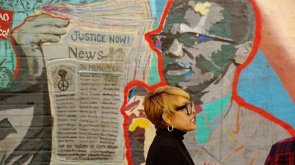 Akemi Kochiyama stands in front of he From Harlem with Love mural of her grandmother Yuri and Black Panther's Malcolm X