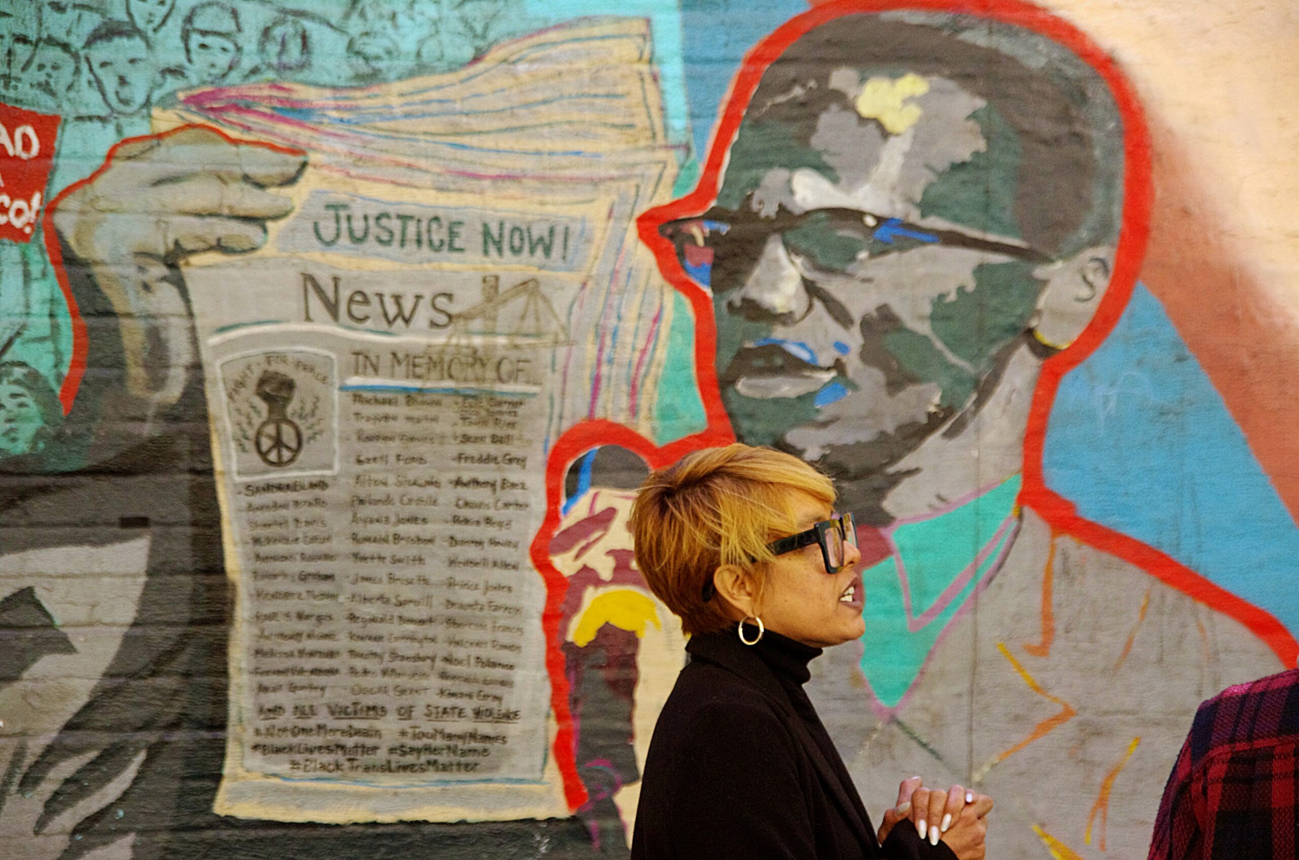 Akemi Kochiyama stands in front of he From Harlem with Love mural of her grandmother Yuri and Black Panther's Malcolm X