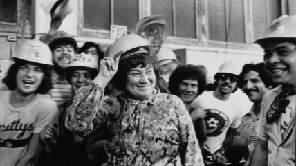 Bella Abzug wearing a hard hat surrounded by a bunch of people also wearing hard hats.