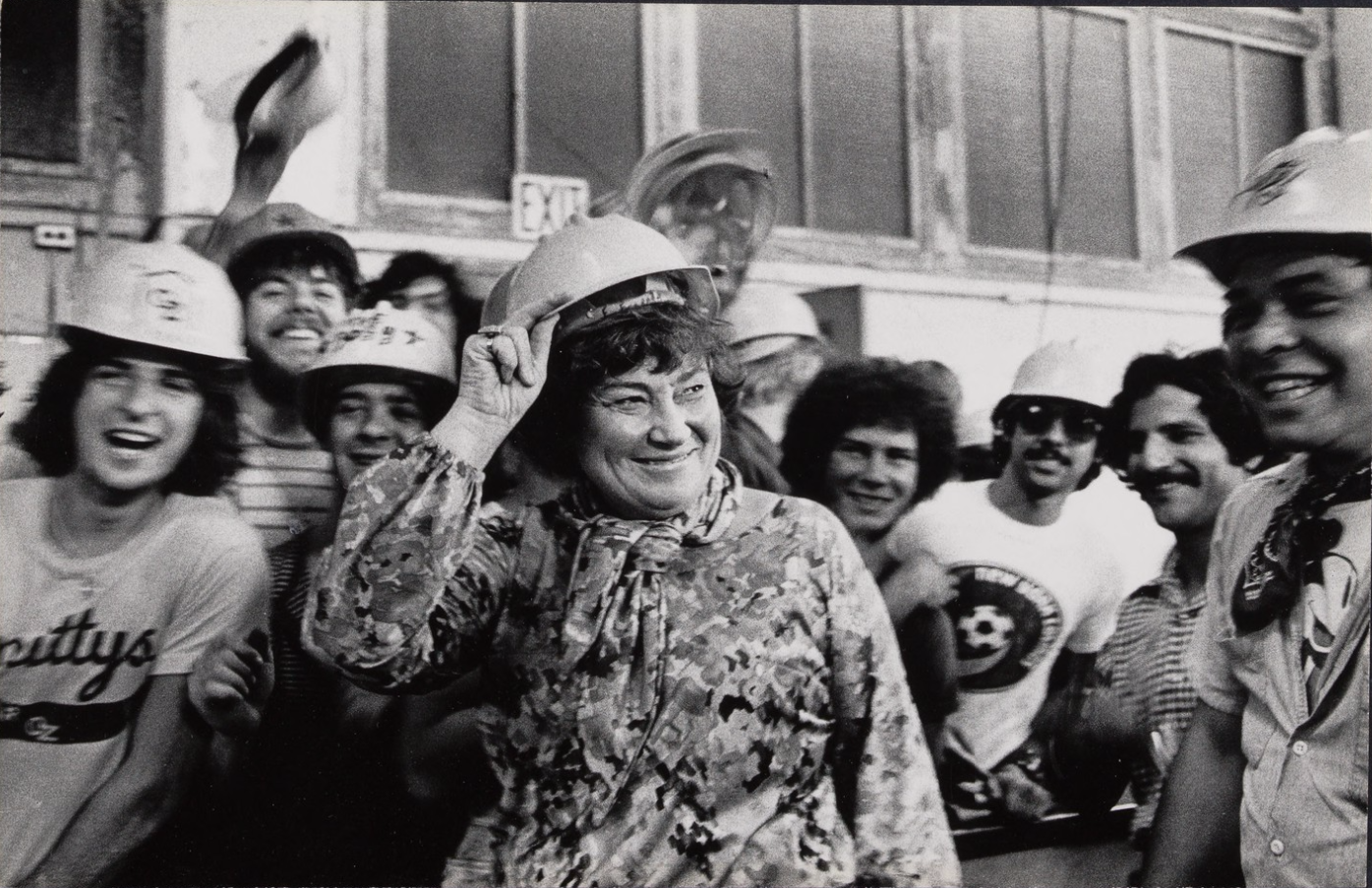 Bella Abzug wearing a hard hat surrounded by a bunch of people also wearing hard hats.