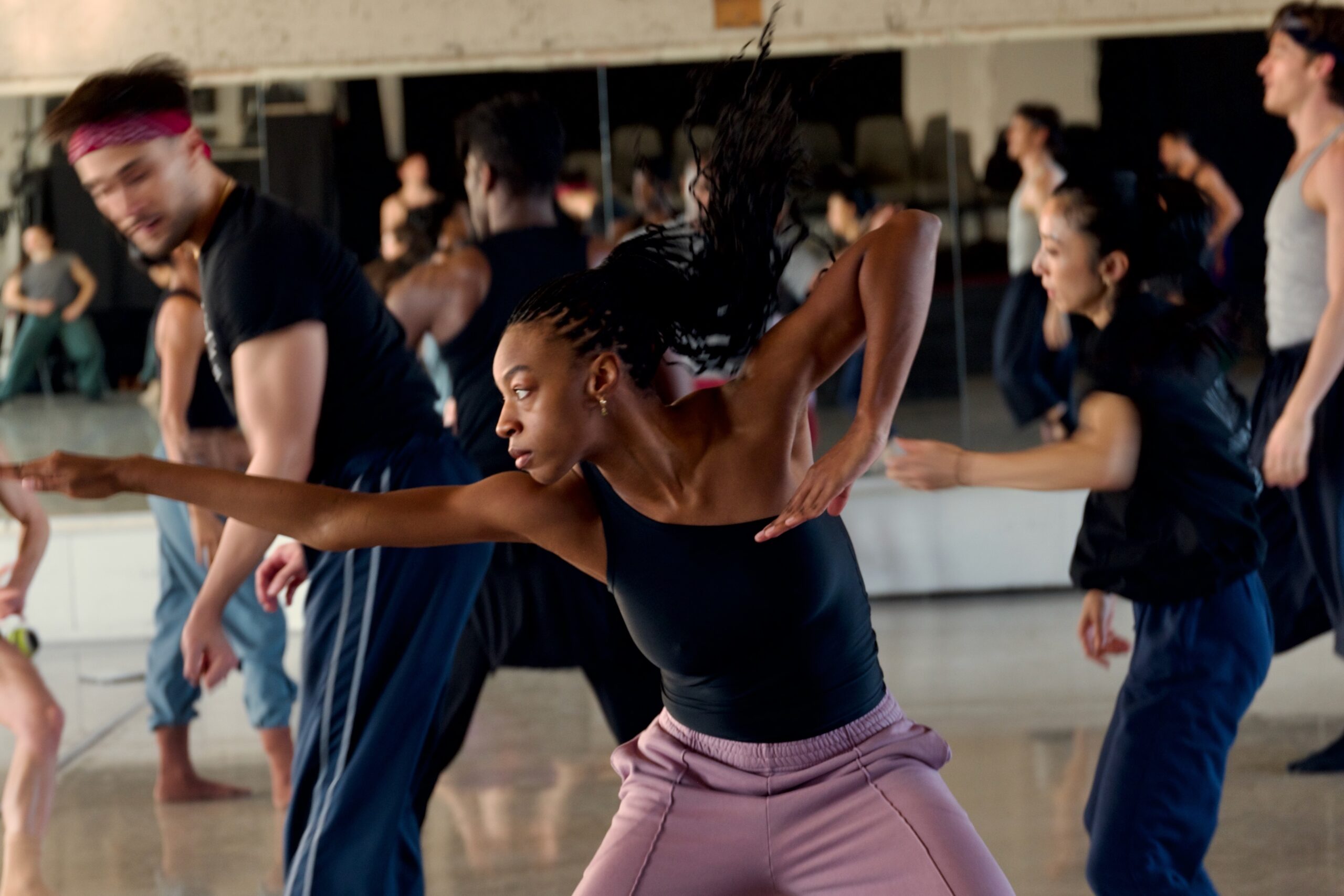 A group of dancers rehearses in a studio with mirrored walls. One dancer in the foreground lunges forward with an extended arm, while others move dynamically behind them.