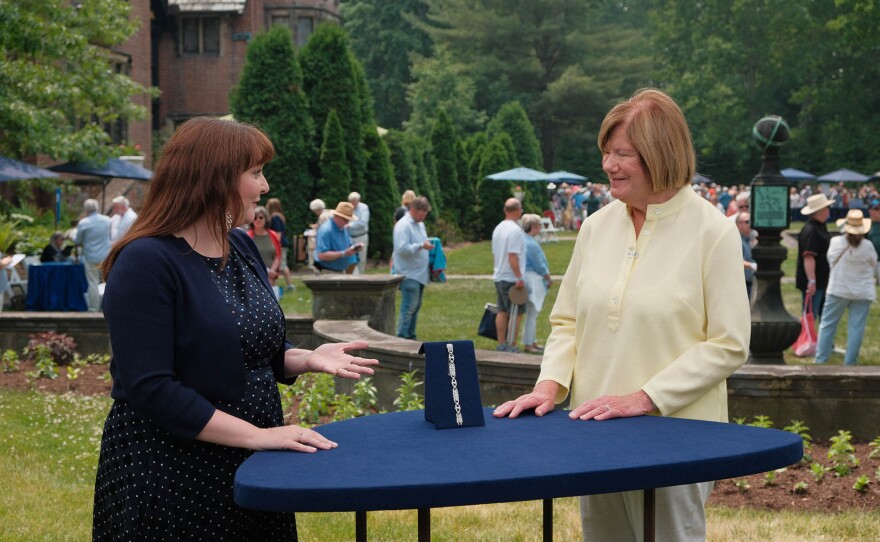 Two women stand on either side of a tall, blue-covered table outdoors. A necklace displayed upright on a small stand sits at the center of the table. In the background, a landscaped garden with trees, stone features, and several people milling about suggests a public outdoor event or gathering.