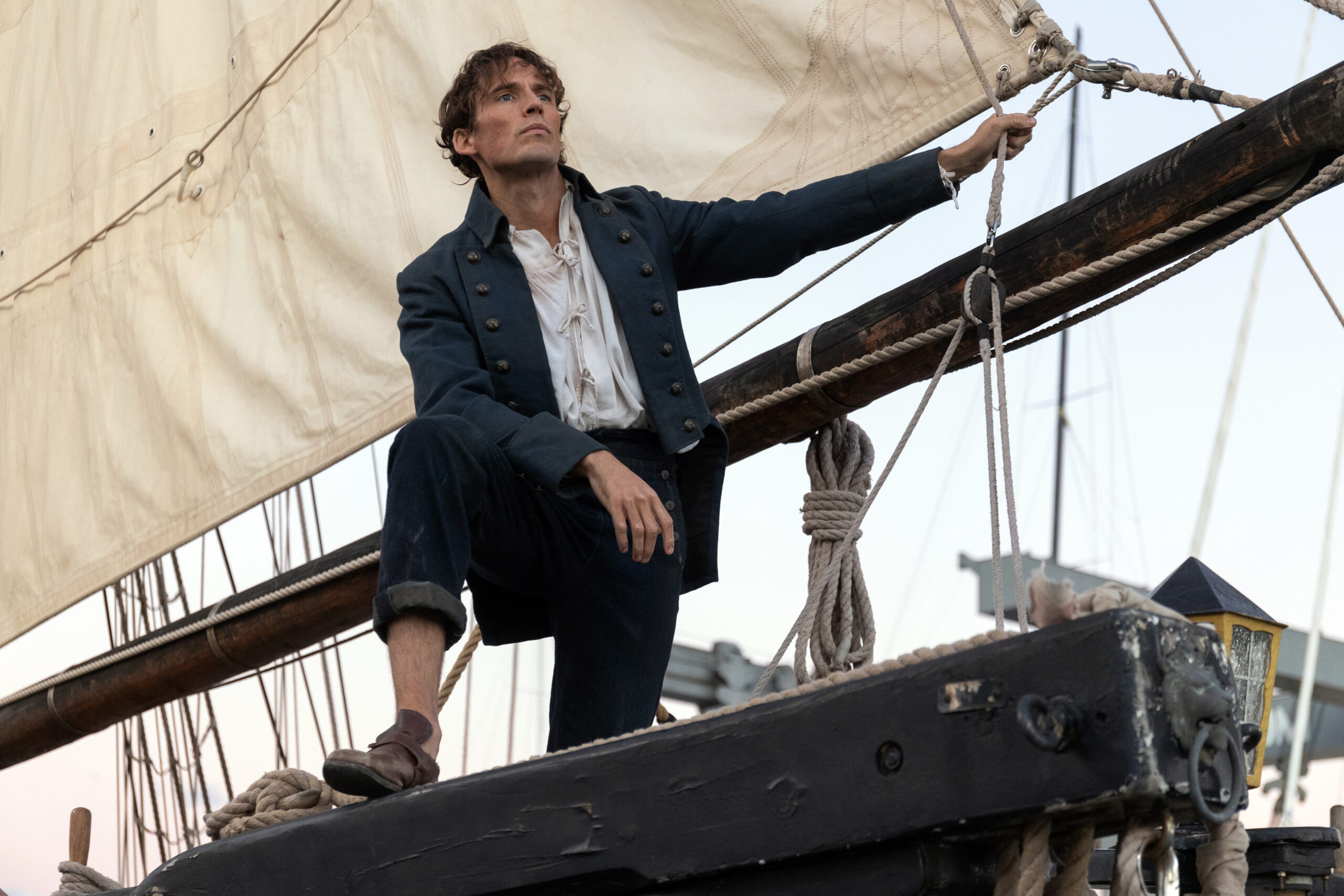 A white man in a blue coat and pants rolled up to his ankles with brown shoes in standing on a ship with a crean colored sail behind him. He is holding on the rope at the mast.