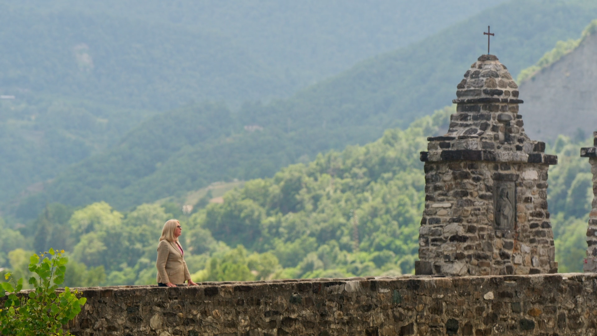 A white woman with blonde hair wearing a tweed blazer is walking up to a brick monument with a cross on top. Behind her is a green mountainous landscape