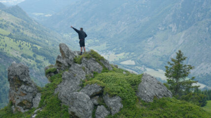 A man wearing a black sweatshirt and shots, stands on top of mountain of rock. He has one hand raised tot he sky. Mountains surround him.