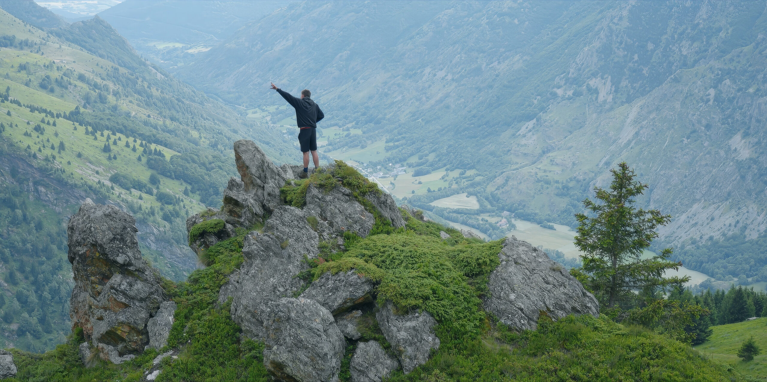 A man wearing a black sweatshirt and shots, stands on top of mountain of rock. He has one hand raised tot he sky. Mountains surround him.
