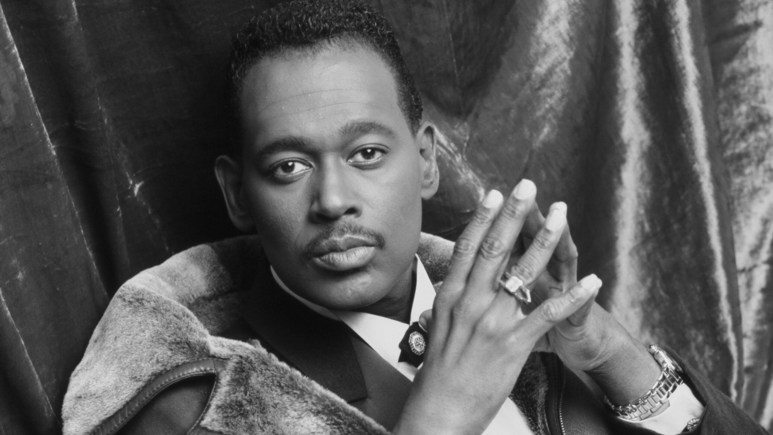 A black and white photo of Luther Vandross wearing a suit, shirt and tie and a leather vest. His hands are crossed, held near his face. One hand has a ring on it.