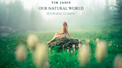 A white woman with long red hair wearing a light brown dress sis on a rock in the middle of a green field with white flowers.