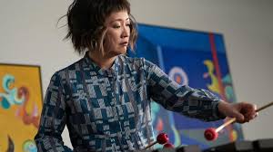 Japanese percussionist Haruka Fujii wearing a patterned light and dark blu eblouse with drum sticks in her hand