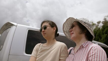 A young asian woman standing next to her mon. the mom is wearing a white floppy hat sun glasses and a pink shirt. The daughter has her black hair pulled back waring a tan t=shirt