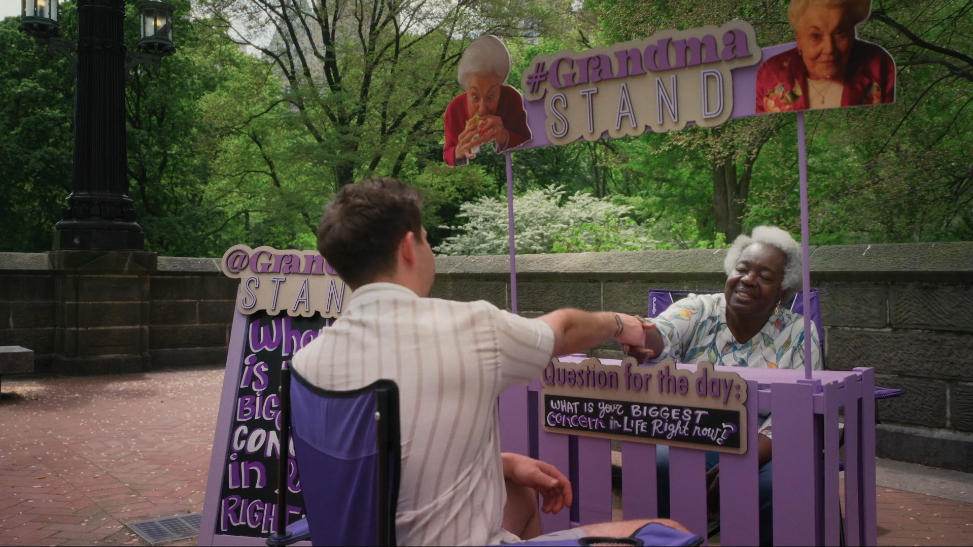 A Black woman with gray hair wearing a green and whote blouse sits at a purple lemonade stand looking desk that says Grandma Stand." She is shaking the had of a 28 year old white man.