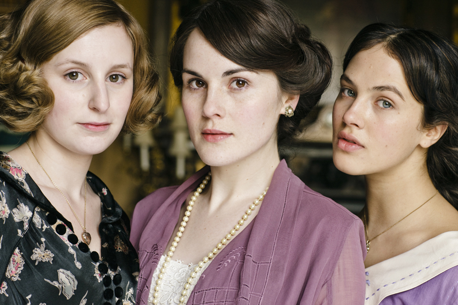 Three white woman pose closely together in a softly lit indoor setting, facing the camera. Each person is framed from the shoulders up and wears early-20th-century-style clothing, including patterned fabric, a lavender outer garment with a pearl necklace, and a light dress with a delicate neckline. The background is softly blurred, emphasizing the trio’s detailed hairstyles, vintage attire, and composed expressions.