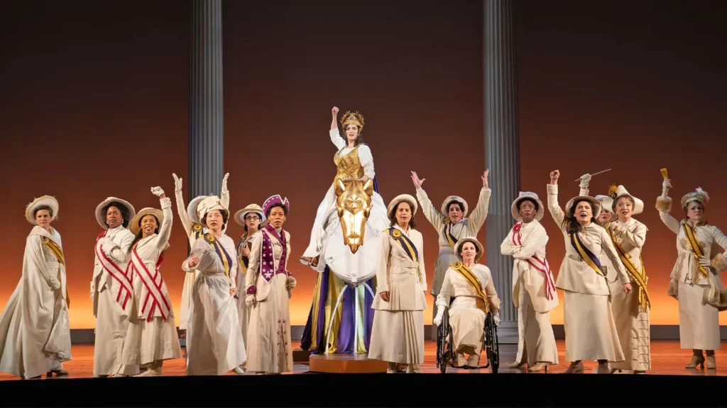 A group of women part of the suffragists wearing a shite suits and sashes stand on stage holding one arm up.