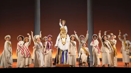 A group of women part of the suffragists wearing a shite suits and sashes stand on stage holding one arm up.