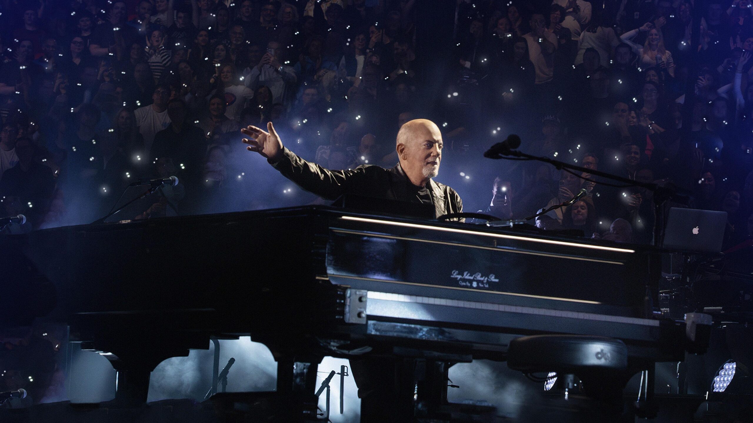 Billy Joel waring a black shirt standing behind a piano on sage with a crowd in the background holding lit flash lighs