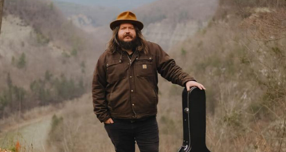 A white man with a brown beard and long hair, wearing a brown hat, jacket and pants leaning on a guitar in the middle of a forest.