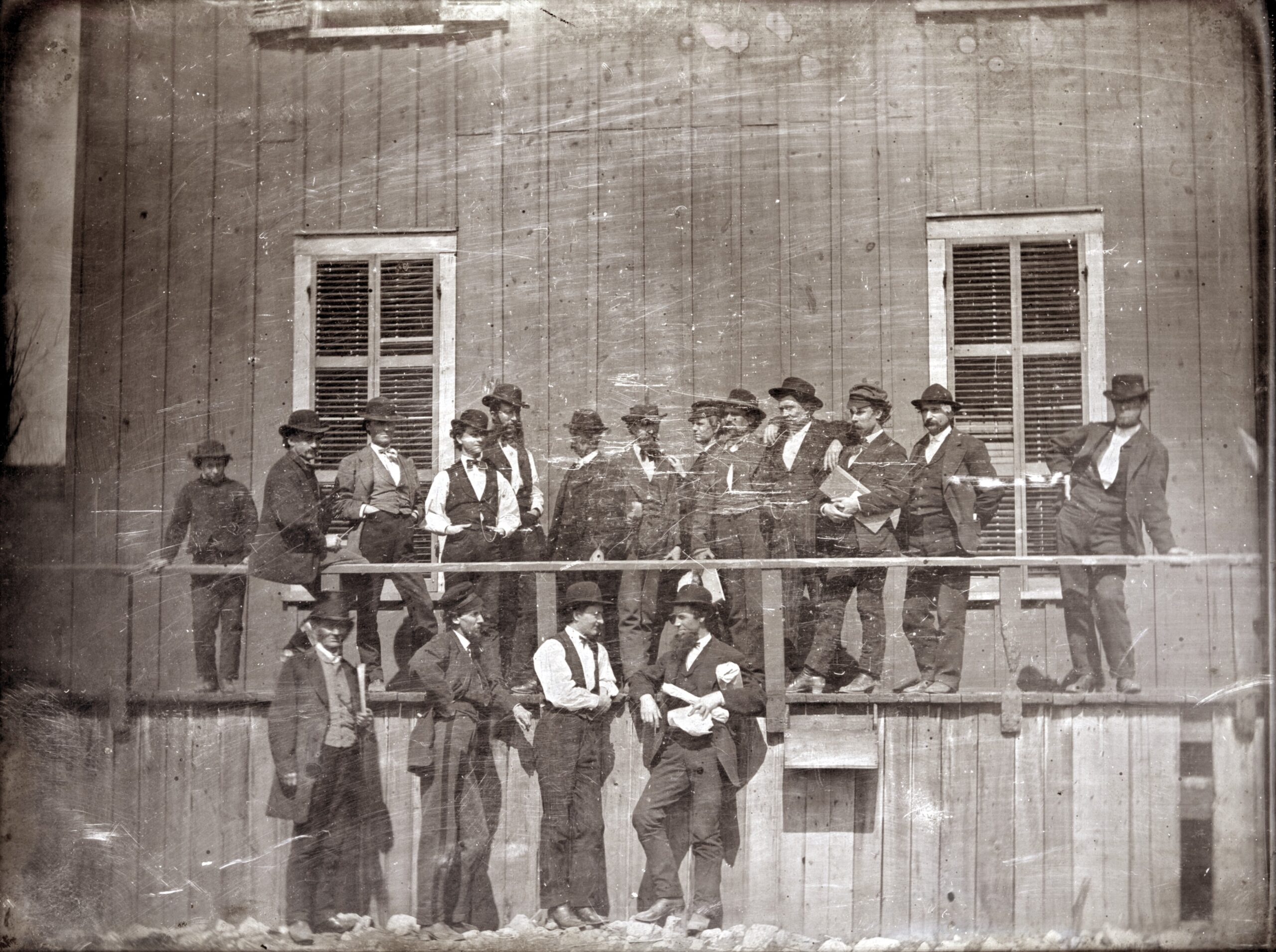 Archival photo of wooden building with a raised platform and a group of men in suits are standing the platform and on the ground.