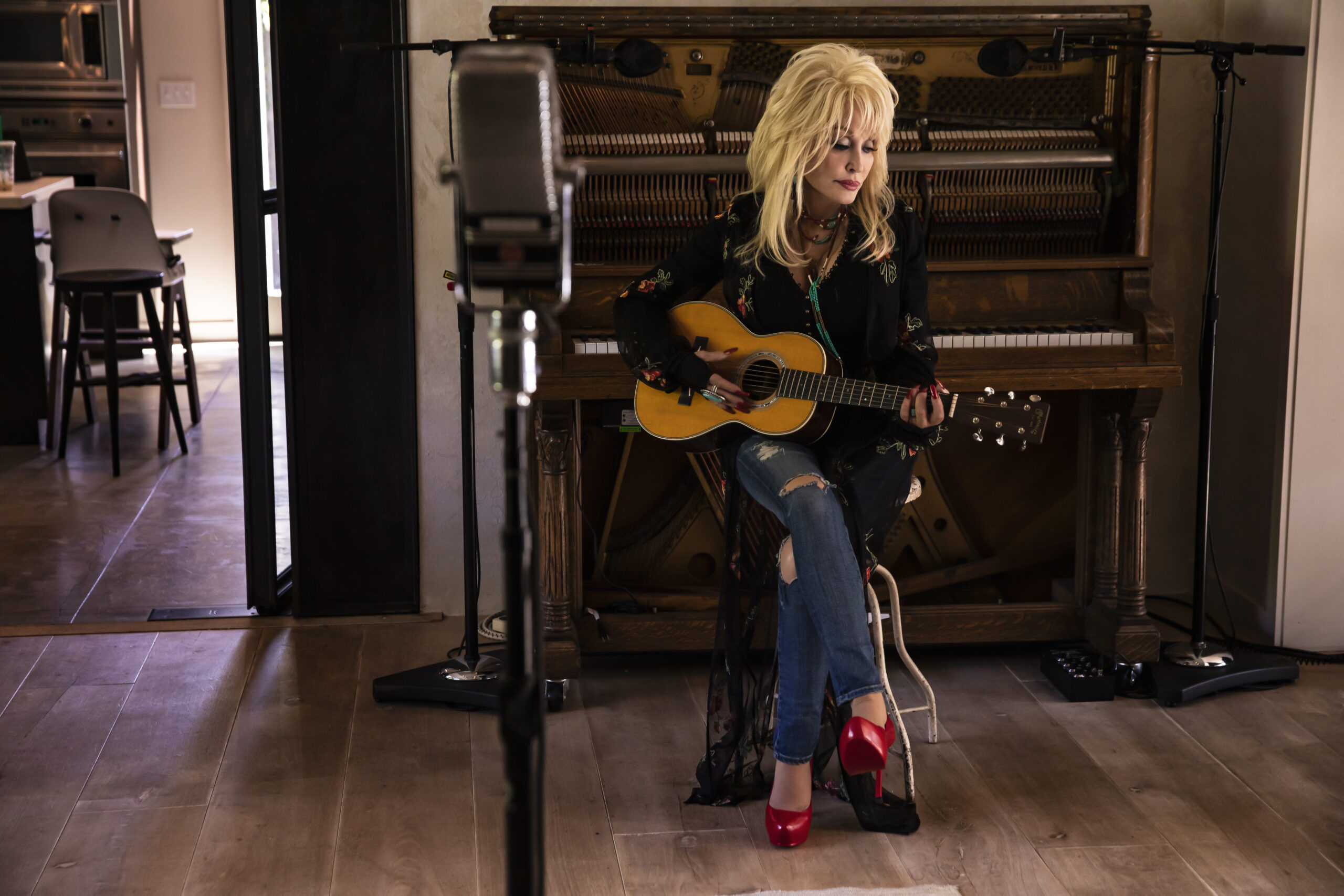 Dolly Parton sitting in front of an upright piano with a guitar in her hand and a microphone ahead of her.