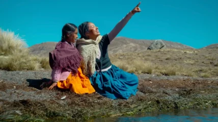 A young Latina girl sits with Grandmother in an open field. The grandma is pointing up at the sky.