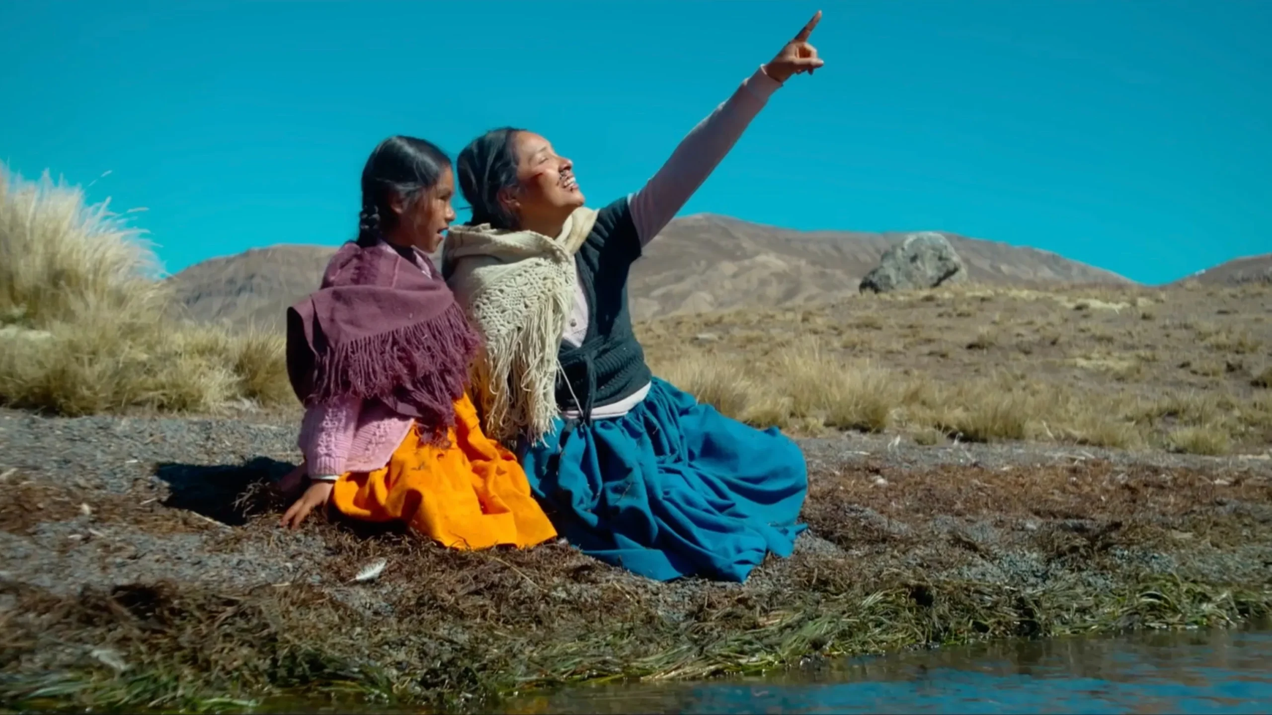 A young Latina girl sits with Grandmother in an open field. The grandma is pointing up at the sky.